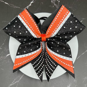 May include: A black and orange cheerleading bow with white accents and sparkling rhinestones. The bow is made of glittery material and has a black elastic band. The bow is on a white surface.