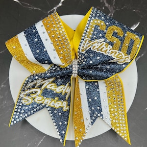 Op de afbeelding: Een marineblauwe, gouden en witte glitter cheerleading strik met de tekst "PSD Vikings" en "Lady Senior" in zilveren strass. De strik heeft een gele midden en is versierd met talrijke strass.