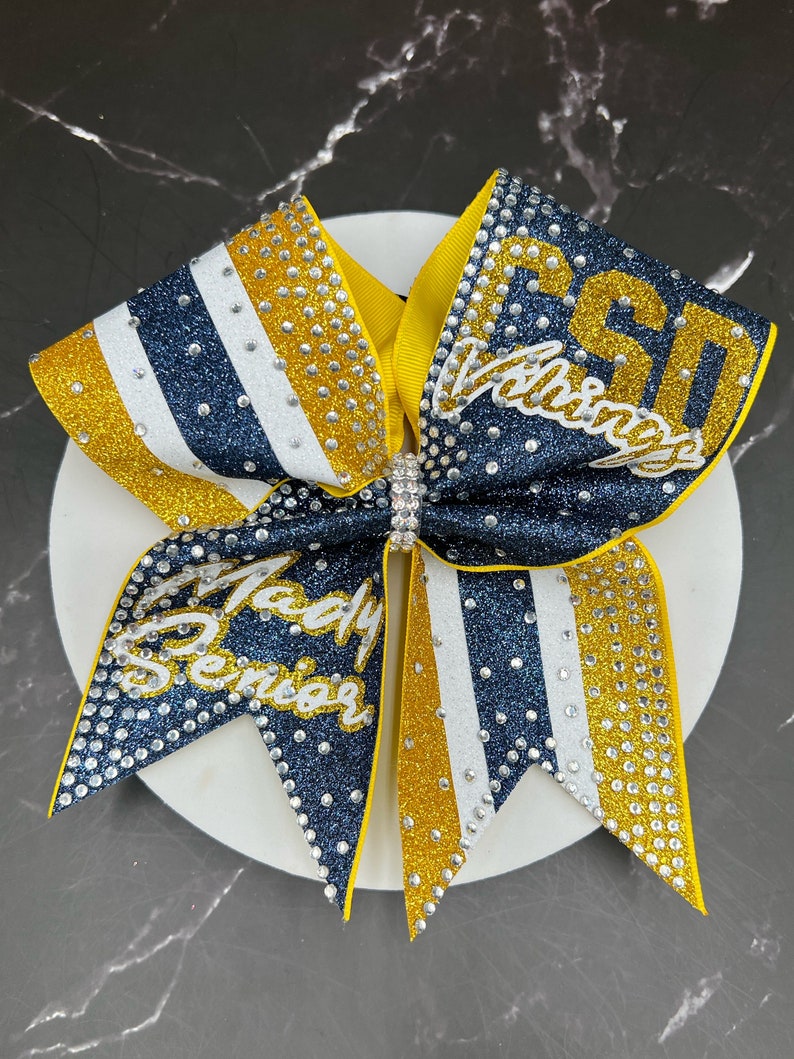 Op de afbeelding: Een marineblauwe, gouden en witte glitter cheerleading strik met de tekst "PSD Vikings" en "Lady Senior" in zilveren strass. De strik heeft een gele midden en is versierd met talrijke strass.