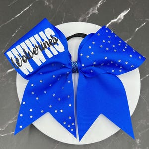 May include: A bright blue hair bow featuring a black elastic band, a center knot with blue glitter, and clear rhinestones scattered across the fabric. The left loop displays the word Wolverines in black script over a white block background.