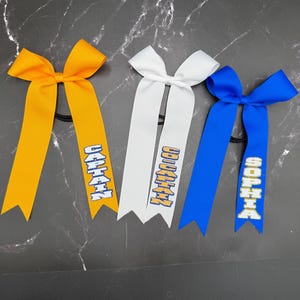 May include: Three hair bows in yellow, white, and blue. Each bow has a ribbon with text in a contrasting color. The yellow bow reads "CAPTAIN", the white bow reads "CAPTAIN", and the blue bow reads "SOPHIA".