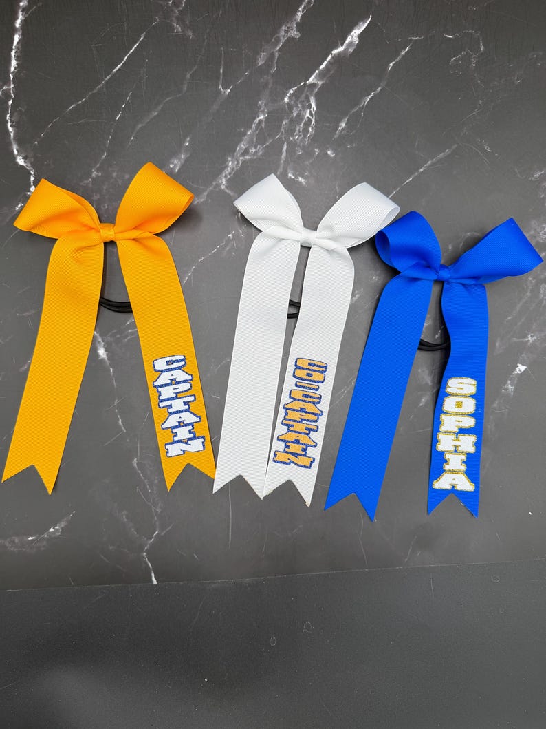 May include: Three hair bows in yellow, white, and blue. Each bow has a ribbon with text in a contrasting color. The yellow bow reads "CAPTAIN", the white bow reads "CAPTAIN", and the blue bow reads "SOPHIA".