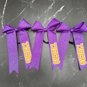 May include: Four purple glitter hair bows with the word "TIGERS" printed in yellow and orange. The bows are on black elastic bands, perfect for cheerleading or team spirit.
