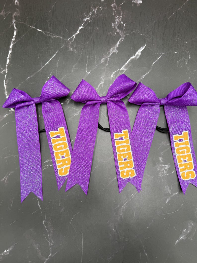 May include: Four purple glitter hair bows with the word "TIGERS" printed in yellow and orange. The bows are on black elastic bands, perfect for cheerleading or team spirit.