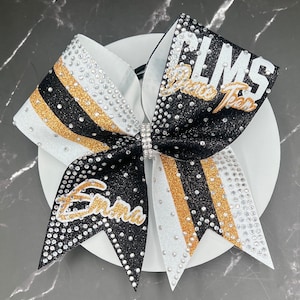 Op de afbeelding: Een cheerleading strik met zwarte, witte en gouden strepen, versierd met strass. De strik heeft de tekst "CLMS Dance Team" en de naam "Emma" in gouden letters. Een strass sluiting zet de strik vast.
