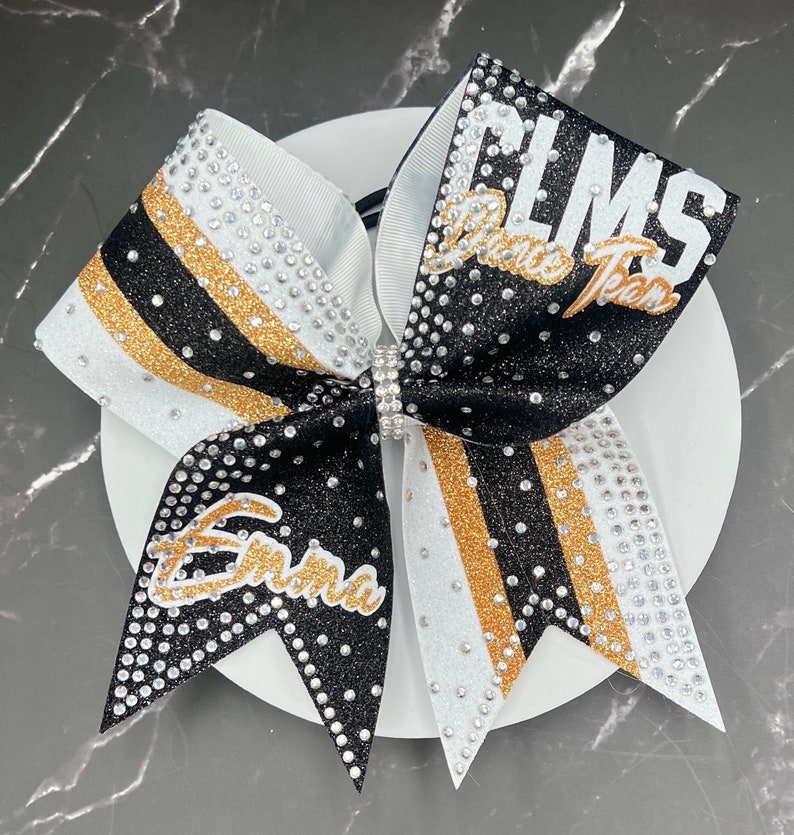 Op de afbeelding: Een cheerleading strik met zwarte, witte en gouden strepen, versierd met strass. De strik heeft de tekst "CLMS Dance Team" en de naam "Emma" in gouden letters. Een strass sluiting zet de strik vast.