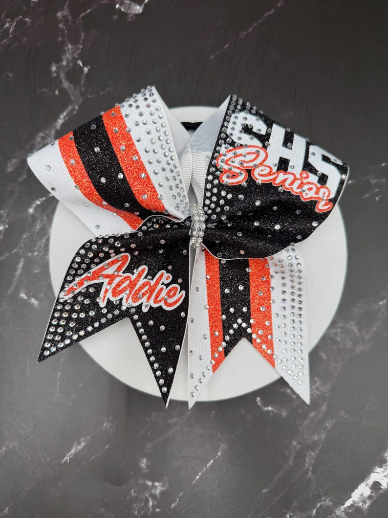 Op de afbeelding: Een cheerleading strik met een wit, zwart en oranje ontwerp. De strik heeft de naam "Addie" in rode letters en de tekst "CHS Senior". Versierd met strass-steentjes, is het een decoratief accessoire.