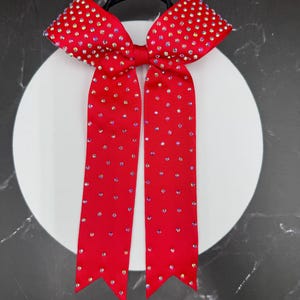 May include: A bright red hair bow made of grosgrain ribbon, featuring two long tails with pointed ends. The entire surface of the bow and its tails is densely decorated with small, iridescent rhinestones that catch the light.