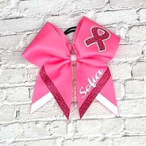 May include: Pink cheerleading bow with a white and glittery red ribbon design. The bow features the name "Sofia" in white script and a red ribbon with a white outline. The bow is attached to a black elastic band.