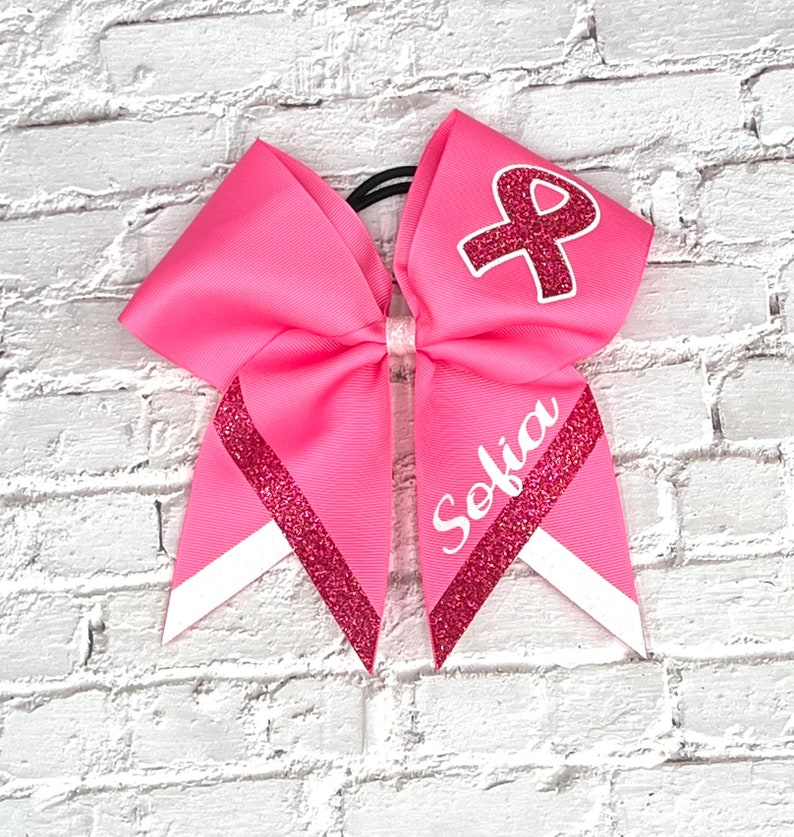 May include: Pink cheerleading bow with a white and glittery red ribbon design. The bow features the name "Sofia" in white script and a red ribbon with a white outline. The bow is attached to a black elastic band.