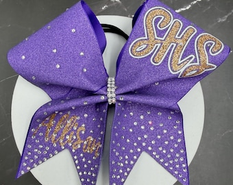 May include: A purple glitter hair bow featuring the letters SHS in gold script on one side and the name Allison in gold script on the other. The bow is embellished with numerous clear rhinestones and a central band of small clear crystals.