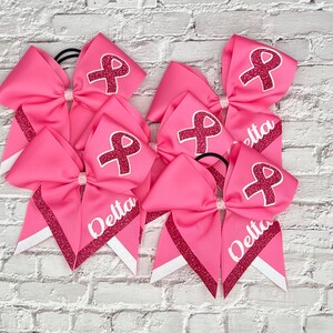 May include: Pink cheerleading bows with a glitter ribbon design and a white ribbon accent. Each bow has a pink ribbon graphic and the word "Delta" in white. The bows are on a white brick background.