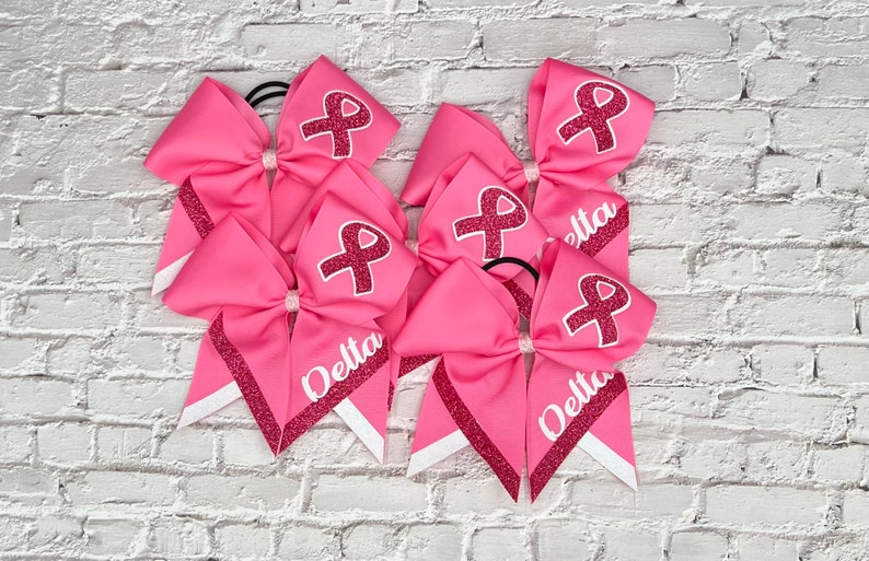 May include: Pink cheerleading bows with a glitter ribbon design and a white ribbon accent. Each bow has a pink ribbon graphic and the word "Delta" in white. The bows are on a white brick background.