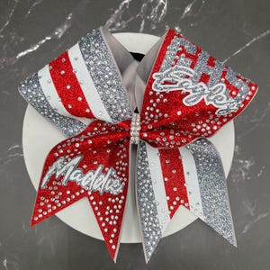May include: A large cheer bow featuring red, white, and silver glitter fabric with scattered clear rhinestones. The bow displays the name Maddie in silver script on one tail and the word Eagles in silver script on the other, with a rhinestone center.