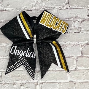 May include: Black glitter cheerleading bow with gold and white stripes. The bow features the name "Angelica" and the word "Wildcats" in gold lettering. A rhinestone accent is in the center.