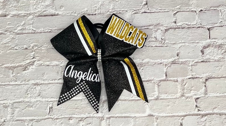 May include: Black glitter cheerleading bow with gold and white stripes. The bow features the name "Angelica" and the word "Wildcats" in gold lettering. A rhinestone accent is in the center.