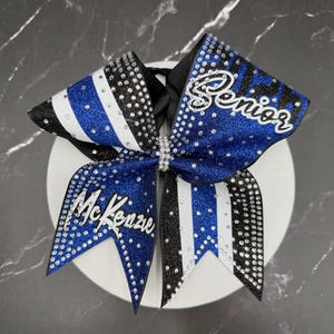 Op de afbeelding: Een blauw, zwart en witte cheerleading strik met de naam "McKenzie" en het woord "Senior" in witte letters. De strik is versierd met strass en heeft een zwart lint in het midden.
