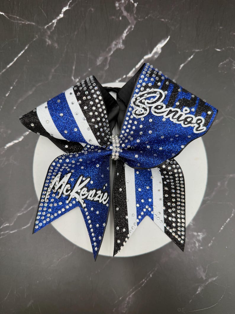 Op de afbeelding: Een blauw, zwart en witte cheerleading strik met de naam "McKenzie" en het woord "Senior" in witte letters. De strik is versierd met strass en heeft een zwart lint in het midden.
