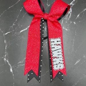 May include: A red glitter hair bow with black ribbon trim and silver rhinestones. The right tail features the word HAWKS in silver lettering outlined with rhinestones. The bow is constructed from layered fabric with a central knot and two long tails.