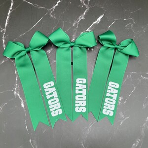 May include: Three green ribbon bows with the word "GATORS" printed in white. The bows are arranged on a dark, marbled surface. The ribbons have a pointed cut at the bottom.