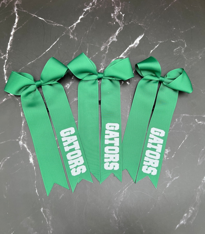 May include: Three green ribbon bows with the word "GATORS" printed in white. The bows are arranged on a dark, marbled surface. The ribbons have a pointed cut at the bottom.