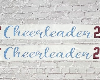 May include: Two white ribbons with text for cheerleaders. The top ribbon says "Alaska Cheerleader 2024" with a maroon megaphone graphic. The bottom ribbon says "Leah Cheerleader 2024" with a maroon megaphone graphic.