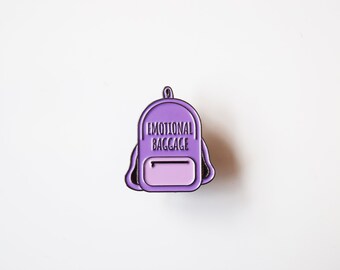 May include: A purple enamel pin in the shape of a backpack. The pin features the words "EMOTIONAL BAGGAGE" in white lettering. The backpack has a lighter purple pocket and a black outline.