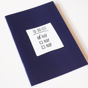 Può includere: Un taccuino blu navy con un adesivo bianco che recita "TO-DO LIST". Il primo elemento, "NAP", è spuntato. Gli altri due elementi della lista sono anch'essi "NAP". Il taccuino è chiuso e appoggiato su una superficie bianca.