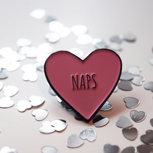 Può includere: Una spilla a forma di cuore in smalto con la scritta "NAPS" in lettere nere. La spilla è di colore rosa intenso con un bordo nero. Coriandoli argentati a forma di cuore circondano la spilla su una superficie rosa chiaro.