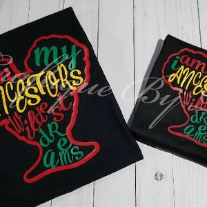 Op de afbeelding: Twee zwarte T-shirts met een rood, groen en geel afro-silhouetontwerp. De tekst op de shirts luidt "Ik ben de wildste dromen van mijn voorouders".
