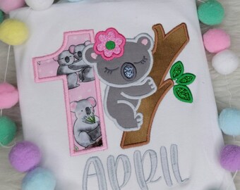 Könnte beinhalten: Ein weißes T-Shirt mit einem gestickten Koala-Design in Rosa und Grau. Der Koala hält eine Blume und sitzt auf einem Baumzweig. Die Zahl "1" ist in Rosa gestickt mit einem Koala-Design im Inneren. Der Text "April" ist in Grau unter dem Koala gestickt.