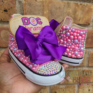 May include: Pink high-top sneakers with a purple bow, embellished with pink and iridescent rhinestones. The sneakers have a white sole and a patch with the text "Doc McStuffins" in gold embroidery.