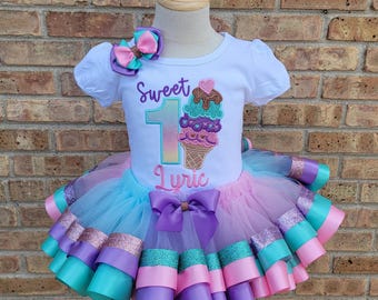 May include: A white short-sleeved shirt with the text "Sweet 1 Lyric" and an embroidered ice cream cone. The shirt is paired with a multi-layered tulle skirt with pink, purple, and teal ribbon accents.