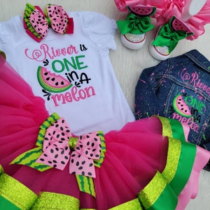 May include: A watermelon-themed outfit set, including a white t-shirt with the text "River is ONE in a melon", a pink tutu skirt, a denim jacket, a pair of shoes, and a hair bow. The outfit features watermelon slice designs and green and pink accents.