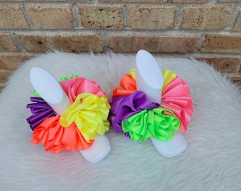May include: Two white hair ties with bright neon pink, yellow, green, and purple satin ribbon ruffles. The hair ties are on a white fluffy surface.