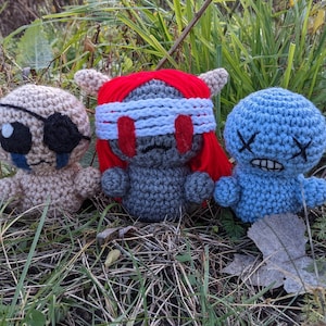 May include: Three crocheted plush toys with different facial expressions. The first toy is tan with a black eye patch, the second is grey with red hair and a white blindfold, and the third is blue with a stitched-on frown.