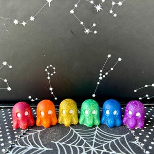 May include: Seven small ghost figurines in a rainbow of colors, from red to purple, arranged in a row on a black and white patterned surface.