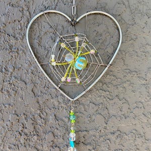 May include: A heart-shaped, silver-toned metal wall hanging. The design features a spider web with a central, colorful bead cluster. Below, a strand of beads in shades of green, blue, and clear hangs from the web. The piece is suspended by a chain.