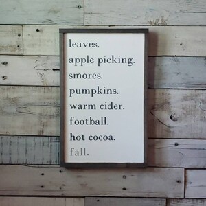 Puede incluir: Un letrero de madera blanco con texto negro que dice "leaves. apple picking. smores. pumpkins. warm cider. football. hot cocoa. fall."