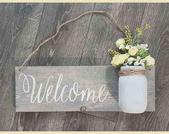 May include: A gray wooden sign with the word "Welcome" written in white paint. A mason jar with yellow flowers is attached to the sign with twine.