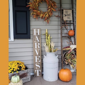 May include: A wooden sign with the word "HARVEST" painted in white letters on a brown background. The sign is leaning against a white wall with a fall-themed wreath and a ladder decorated with pumpkins and fall foliage.