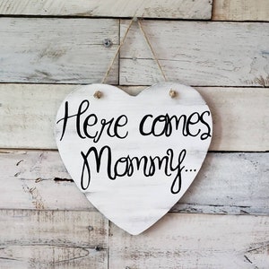 May include: White wooden heart-shaped sign with black calligraphy that reads "Here comes mommy..." hanging on a white wood plank wall.