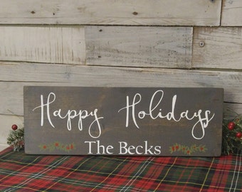 May include: A dark gray wooden sign with white lettering that reads "Happy Holidays The Becks". The sign is decorated with green holly leaves and red berries.