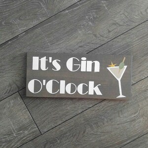 May include: A wooden sign with the text "It's Gin O'Clock" in white lettering. A white martini glass with a green olive and a toothpick is pictured to the right of the text.