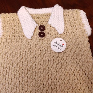 A handmade, beige crochet vest with a white collar and sleeve trim. The vest has two brown buttons and a tag that reads "Handmade with love". The vest is likely for a baby or toddler.