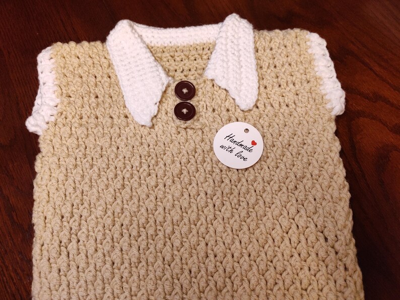 A handmade, beige crochet vest with a white collar and sleeve trim. The vest has two brown buttons and a tag that reads "Handmade with love". The vest is likely for a baby or toddler.