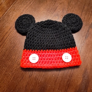 A black and red crocheted hat featuring two round black ears on top and two white four-hole buttons attached to the red band at the bottom. The hat is constructed with a textured yarn stitch pattern throughout the entire piece.