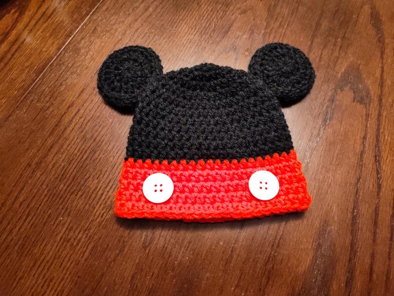A black and red crocheted hat featuring two round black ears on top and two white four-hole buttons attached to the red band at the bottom. The hat is constructed with a textured yarn stitch pattern throughout the entire piece.