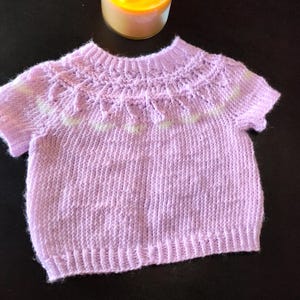A short-sleeved, light purple knitted sweater featuring a round neckline with a textured yoke pattern. The garment includes a ribbed collar, ribbed cuffs, and a ribbed hem, with a subtle white accent line integrated into the yoke design.