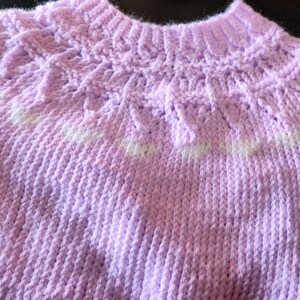 A short-sleeved, light purple knitted sweater featuring a textured yoke with a decorative pattern around the neckline. The garment is constructed with a ribbed collar, ribbed sleeve cuffs, and a ribbed hem, showcasing a consistent knit stitch throughout the body.